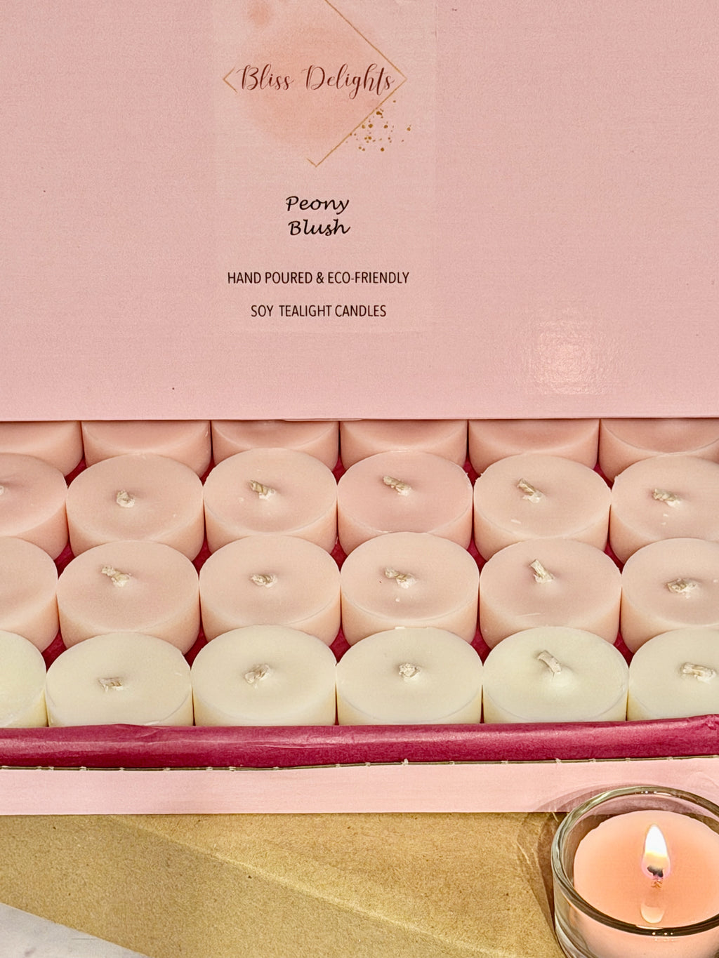 Open box of Bliss Delights Peony Blush soy wax tealights in blush pink and ivory, displayed with pink branded packaging and a lit tealight in a reusable clear glass holder.