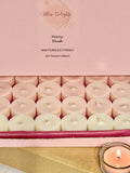 Open box of Bliss Delights Peony Blush soy wax tealights in blush pink and ivory, displayed with pink branded packaging and a lit tealight in a reusable clear glass holder.