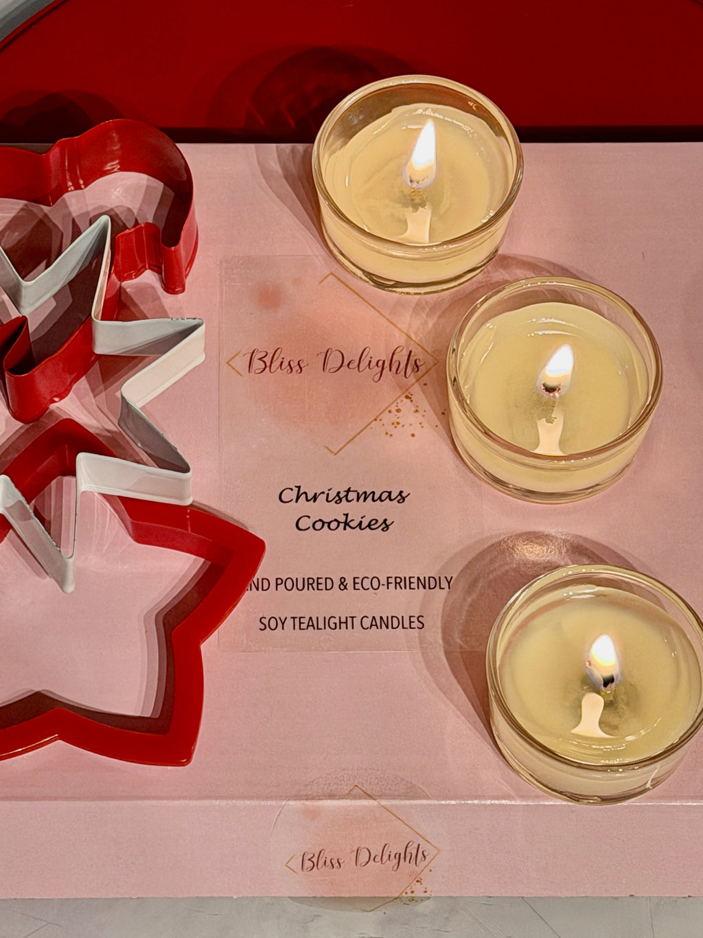 Three lit Bliss Delights Christmas Cookies scented soy tealight candles in clear glass holders, arranged on pink branded packaging alongside red and silver Christmas tree decorations.