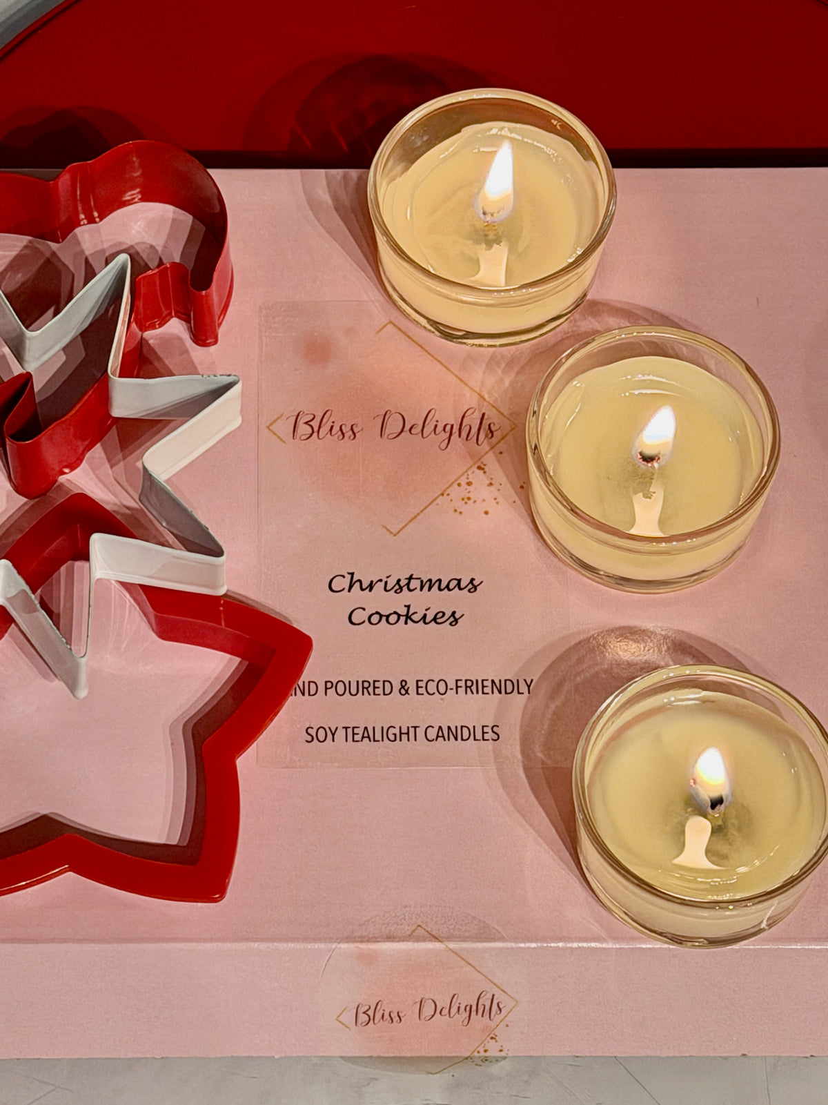 Three lit Bliss Delights Christmas Cookies scented soy tealight candles in clear glass holders, arranged on pink branded packaging alongside red and silver Christmas tree decorations.