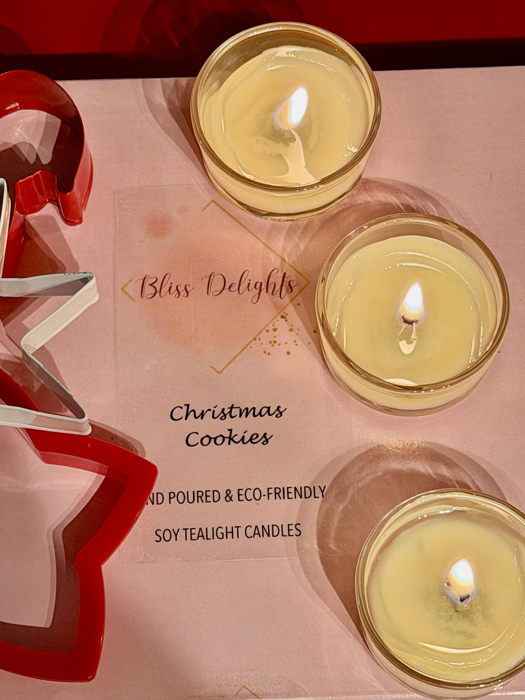 Three lit Bliss Delights Christmas Cookies scented soy tealight candles in clear glass holders, arranged on pink branded packaging alongside red and silver Christmas tree decorations.