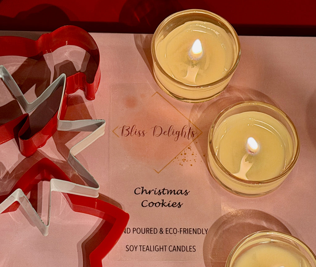 Three lit Bliss Delights Christmas Cookies scented soy tealight candles in clear glass holders, arranged on pink branded packaging alongside red and silver Christmas tree decorations.