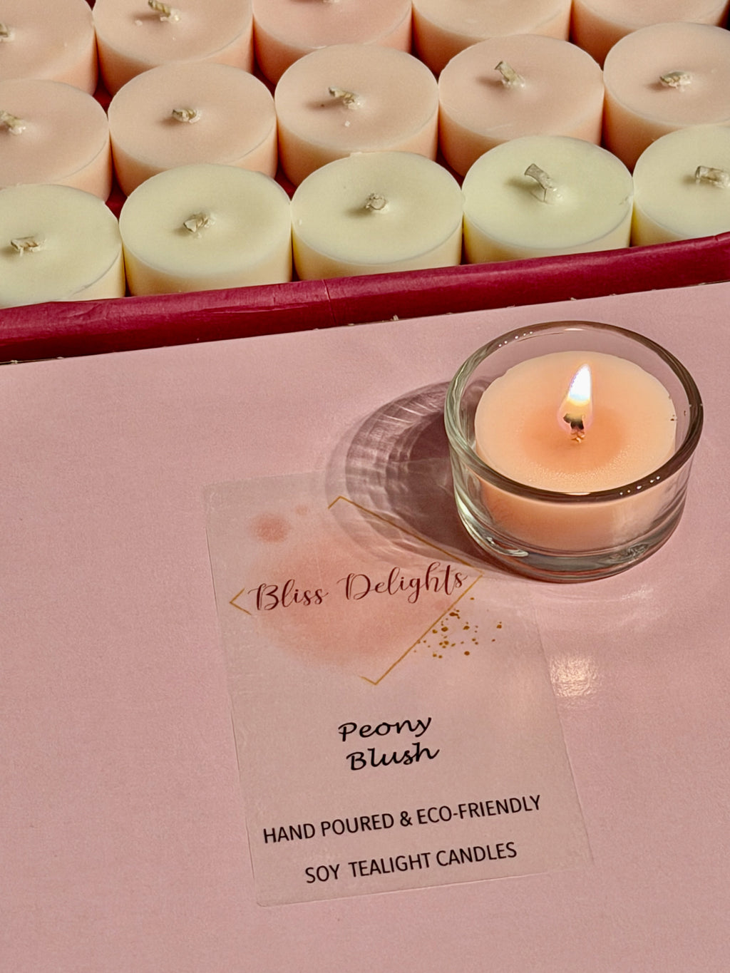 Blush pink Peony Blush soy tealight burning in a clear glass holder on pink Bliss Delights packaging, with pink and ivory tealights displayed in an open box behind it.