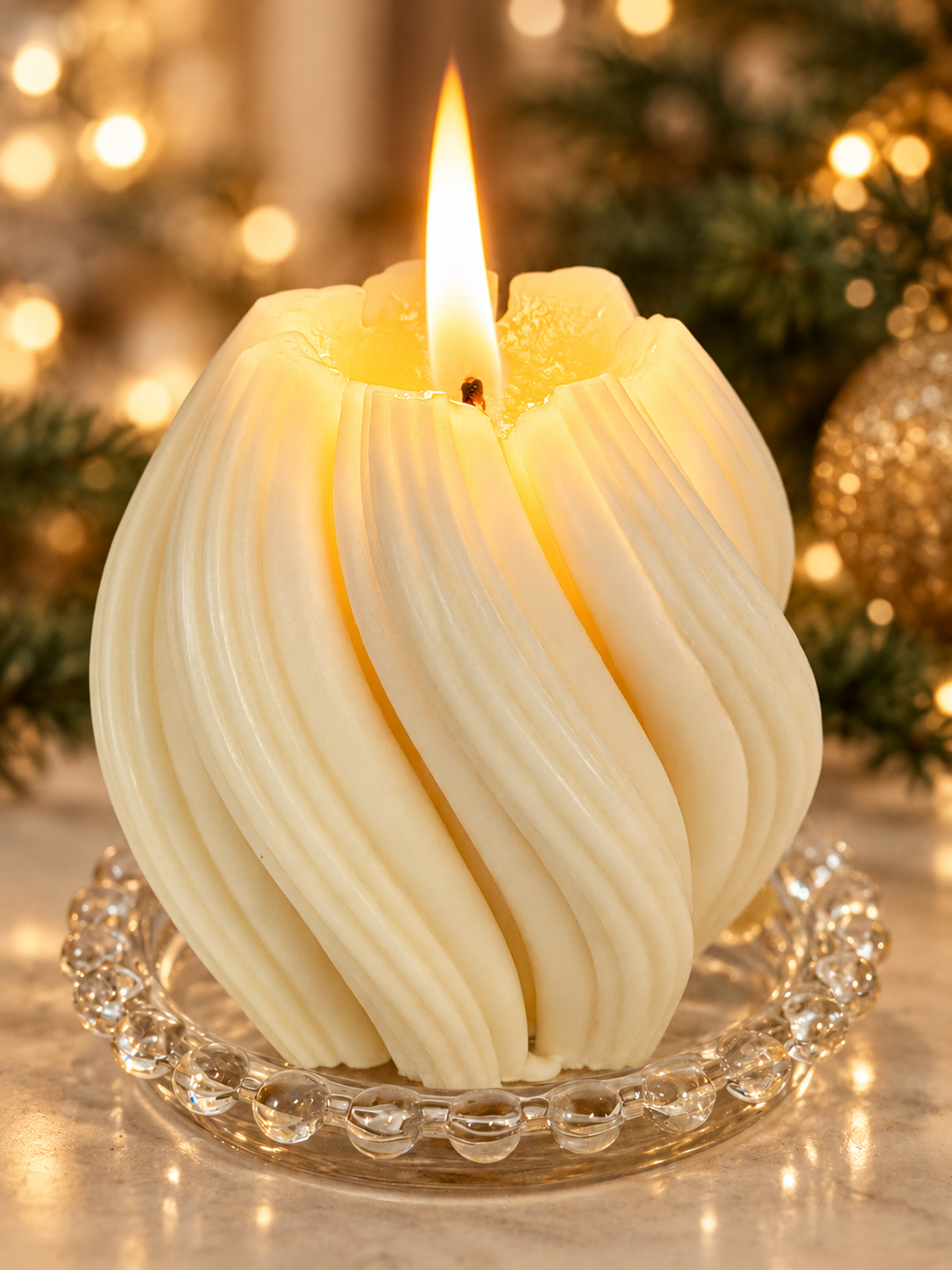 Bliss Delights ivory Twirl soy wax pillar candle with an elegant spiral design, lit on a glass beaded dish against a warm, festive Christmas backdrop with evergreen foliage and golden lights.