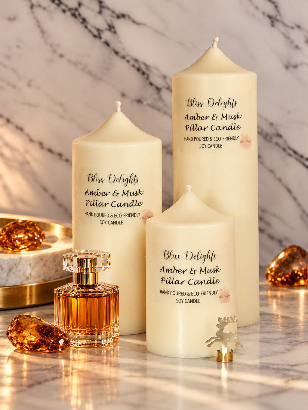 Bliss Delights Amber & Musk set of three ivory soy pillar candles displayed on a marble surface with a small amber perfume bottle, amber crystal accents and a gold reindeer ornament.