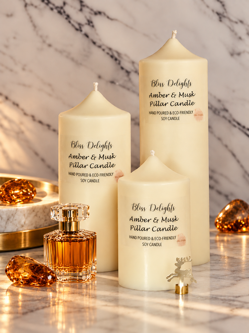 Bliss Delights Amber & Musk set of three ivory soy pillar candles displayed on a marble surface with a small amber perfume bottle, amber crystal accents and a gold reindeer ornament.