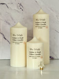 Three cream-coloured Bliss Delights Amber & Musk soy wax pillar candles in small, medium and large sizes, arranged against a white marble background with a decorative gold reindeer ornament.