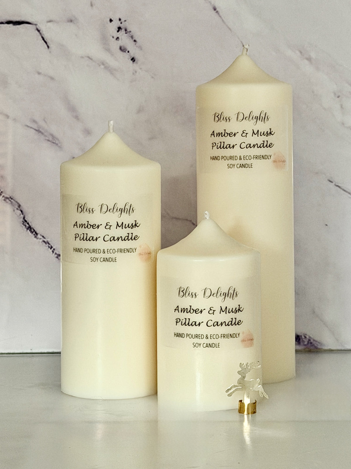 Three cream-coloured Bliss Delights Amber & Musk soy wax pillar candles in small, medium and large sizes, arranged against a white marble background with a decorative gold reindeer ornament.