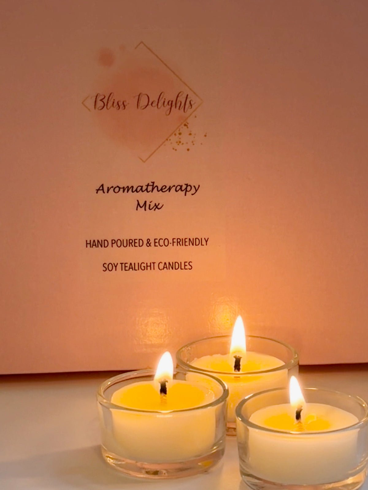 Three glowing Bliss Delights Aromatherapy Mix soy wax tealights burning in reusable clear glass holders in front of pink branded packaging. Hand-poured candles creating a warm, cosy atmosphere.