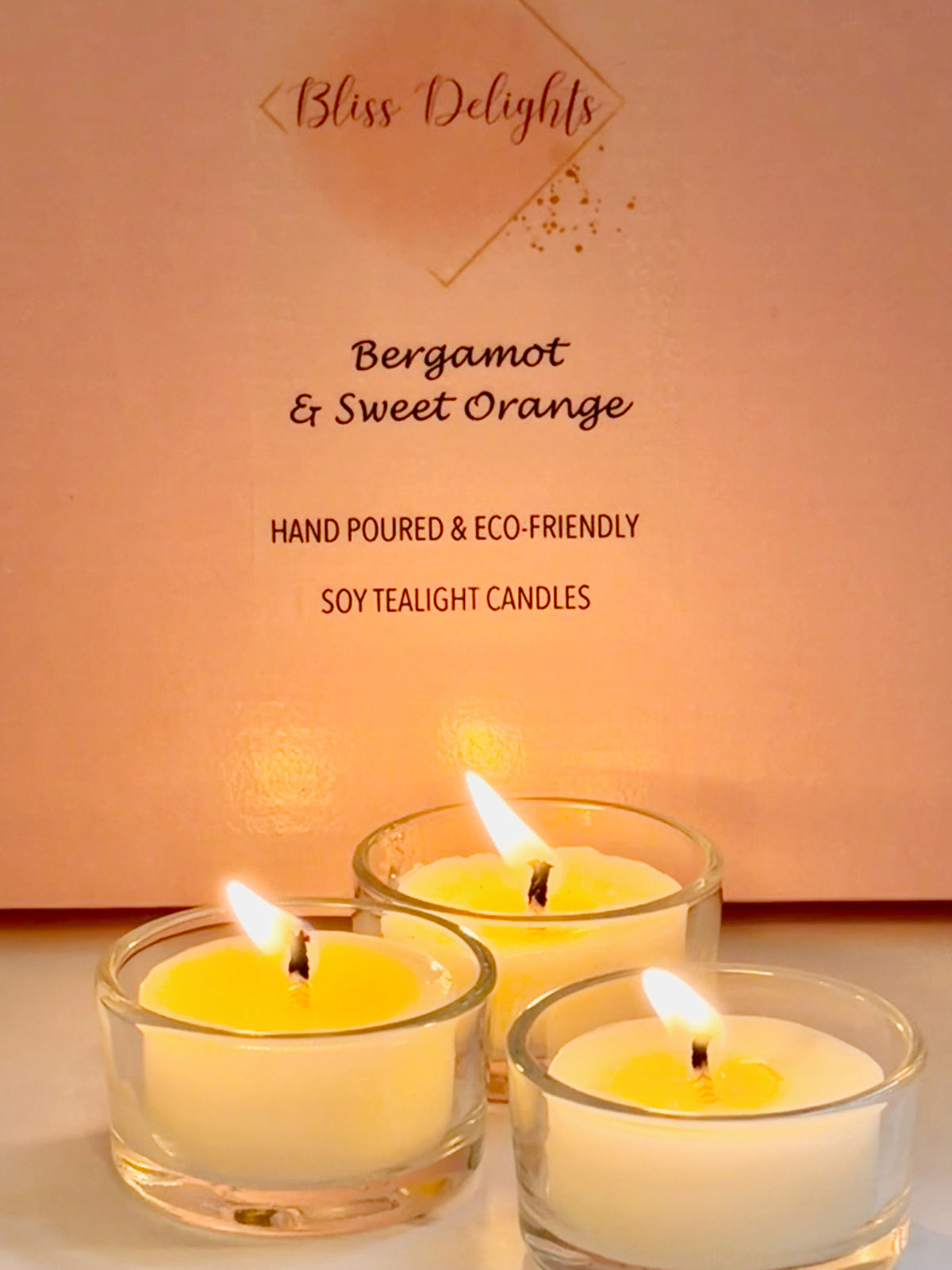 Three glowing Bergamot and Sweet Orange soy wax tealights in reusable clear glass holders, displayed against Bliss Delights pink packaging. Handmade candles with a warm golden glow and a fresh citrus fragrance.
