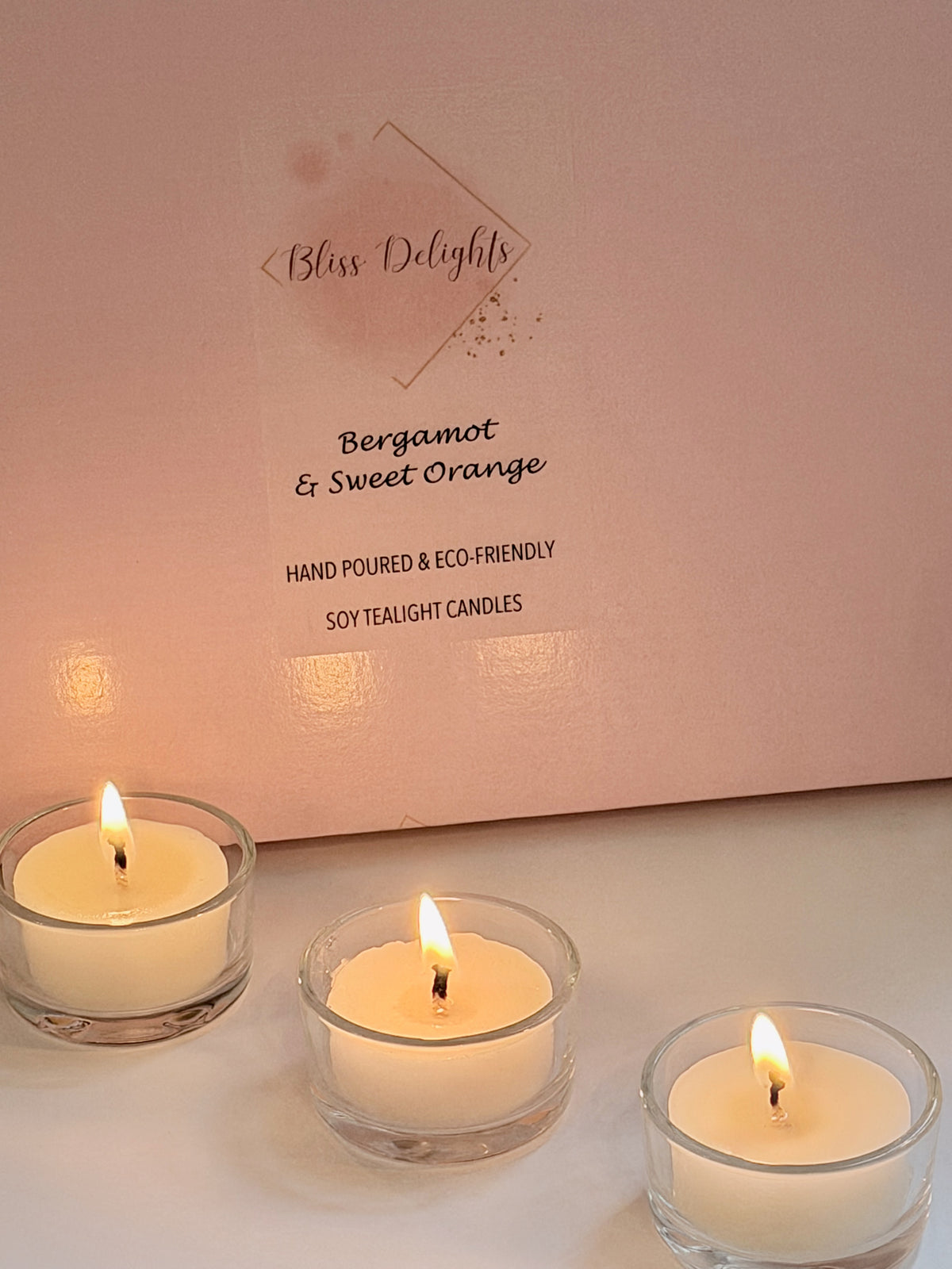 Bliss Delights Bergamot and Sweet Orange soy wax tealights burning in three clear glass holders in front of pink branded packaging. Hand-poured tealight candles creating a warm, inviting glow.