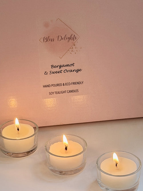 Bliss Delights Bergamot and Sweet Orange soy wax tealights burning in three clear glass holders in front of pink branded packaging. Hand-poured tealight candles creating a warm, inviting glow.