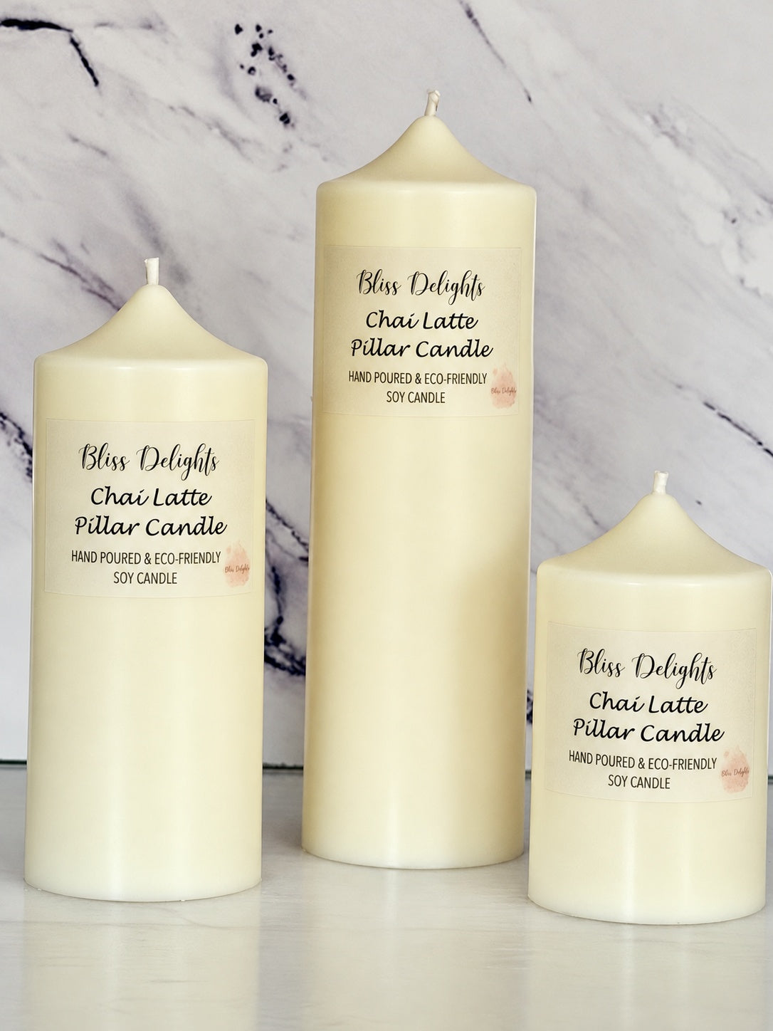 Three cream-coloured Bliss Delights Chai Latte soy wax pillar candles in small, medium and large sizes, arranged on a pale marble surface against a white marble background. Each candle features a Chai Latte label and is hand-poured in the UK.