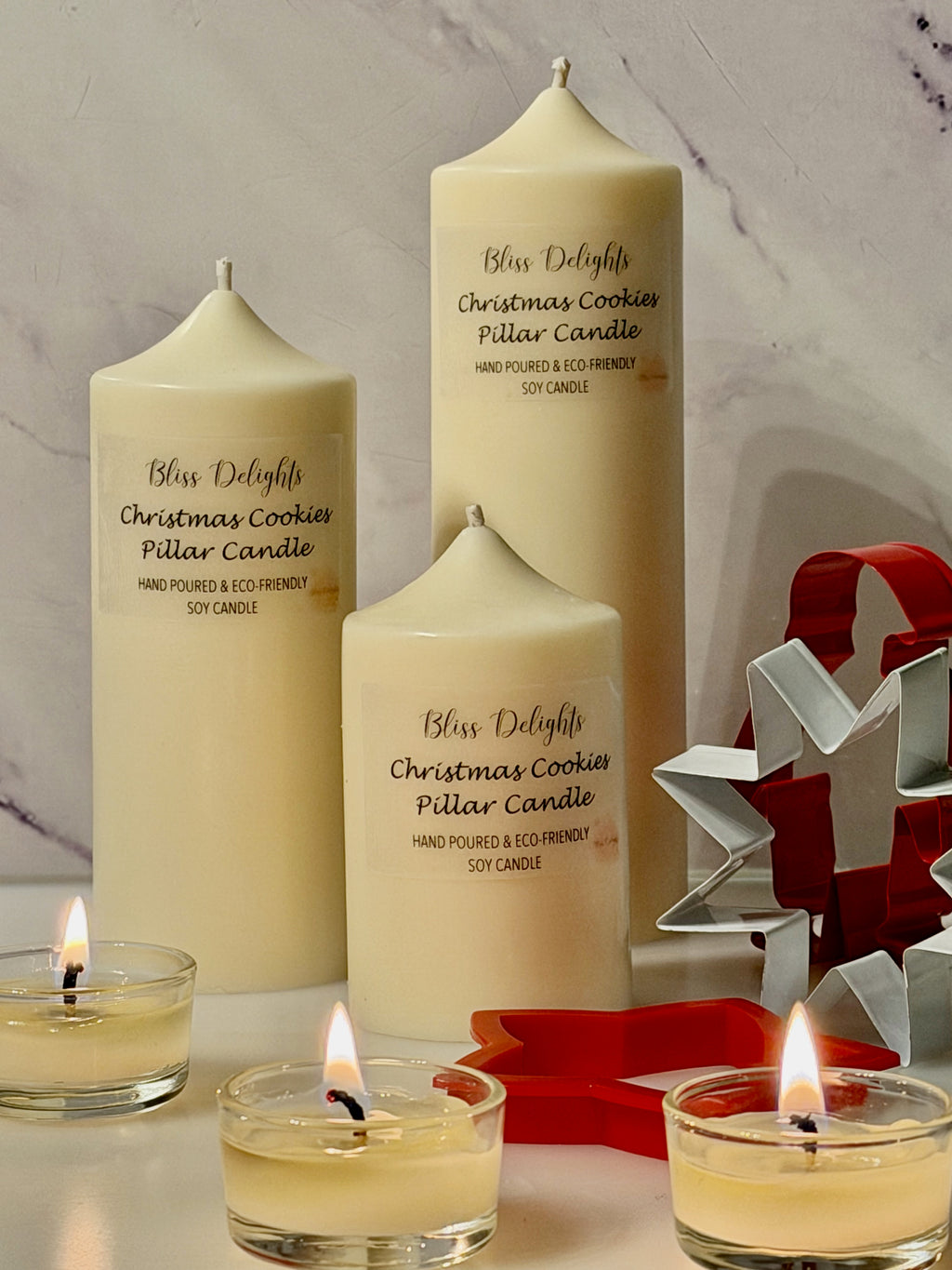 Three cream-coloured Bliss Delights Christmas Cookies soy wax pillar candles in small, medium and large sizes, styled with lit tealight candles and red and silver Christmas tree decorations against a white marble background.