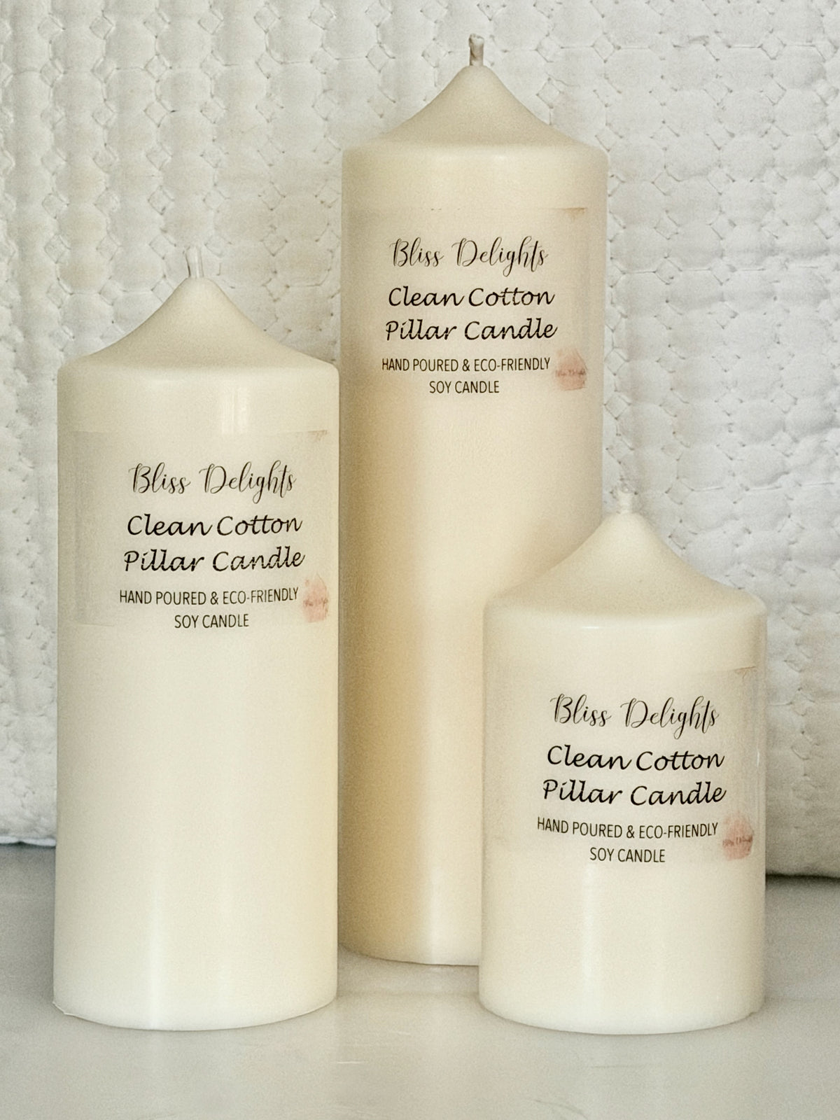 Three cream-coloured Bliss Delights Clean Cotton soy wax pillar candles in small, medium and large sizes, arranged against a white textured quilted background.