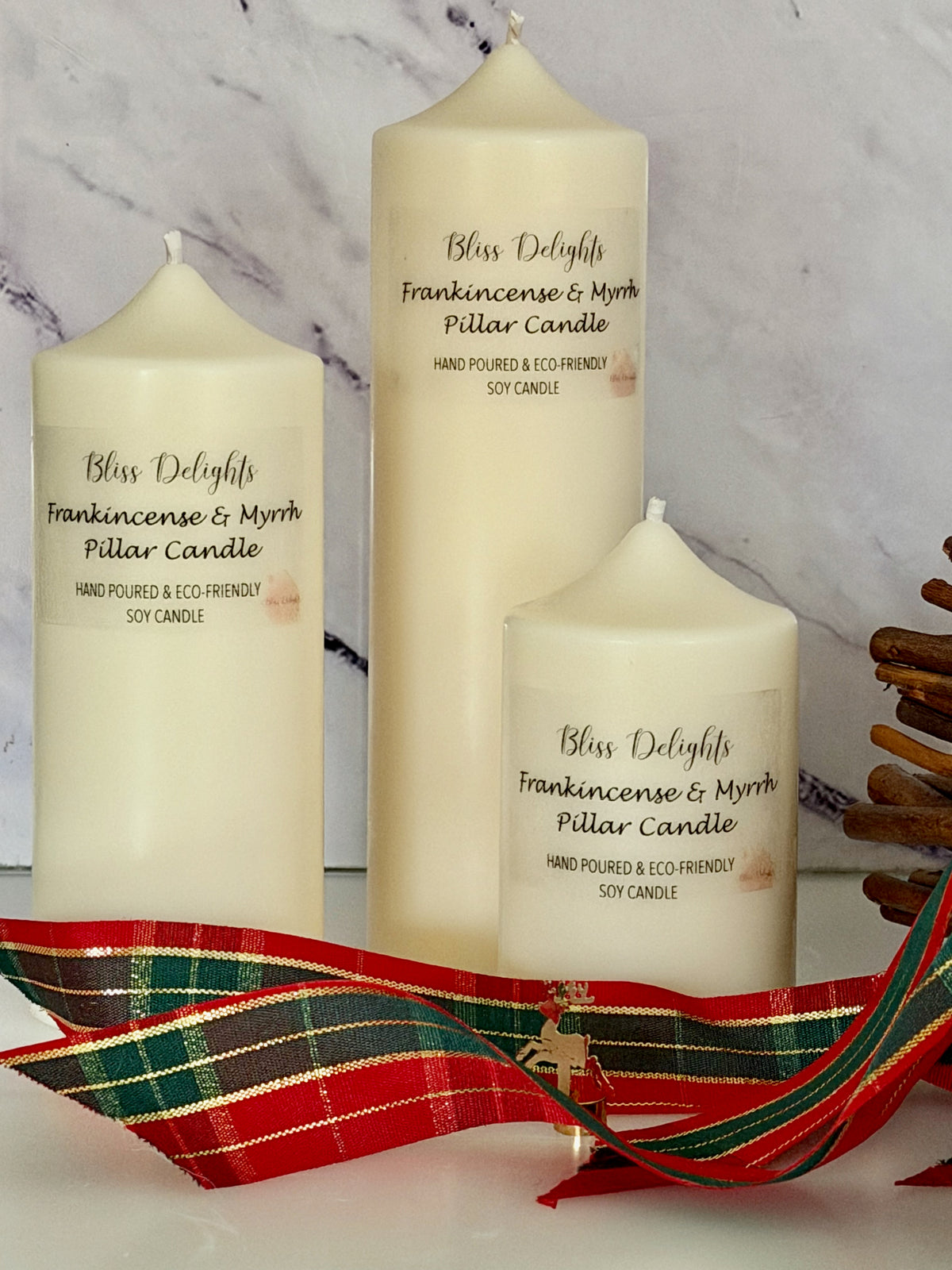 Three cream-coloured Bliss Delights Frankincense & Myrrh soy wax pillar candles in small, medium and large sizes, arranged against a white marble background with festive red and green tartan ribbon and a decorative reindeer ornament.