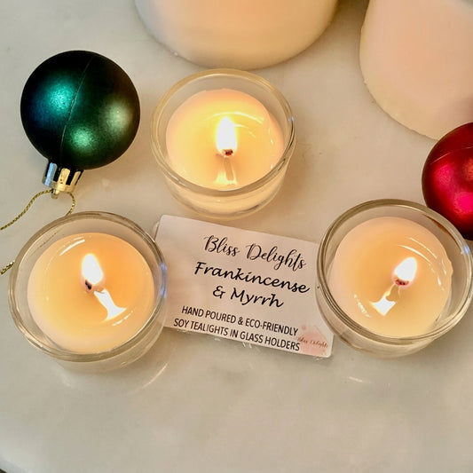 Three lit candles in glass holders scented with Bliss Delights Frankincense & Myrrh on a white surface with Christmas ornaments.