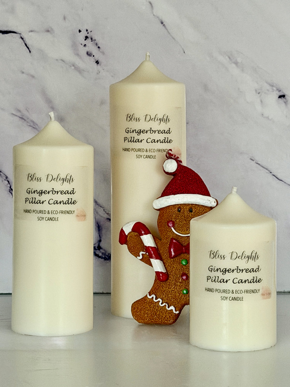 Three cream-coloured Bliss Delights Gingerbread soy wax pillar candles in small, medium and large sizes, arranged with a festive gingerbread figure wearing a Santa hat and holding a candy cane, against a white marble background.