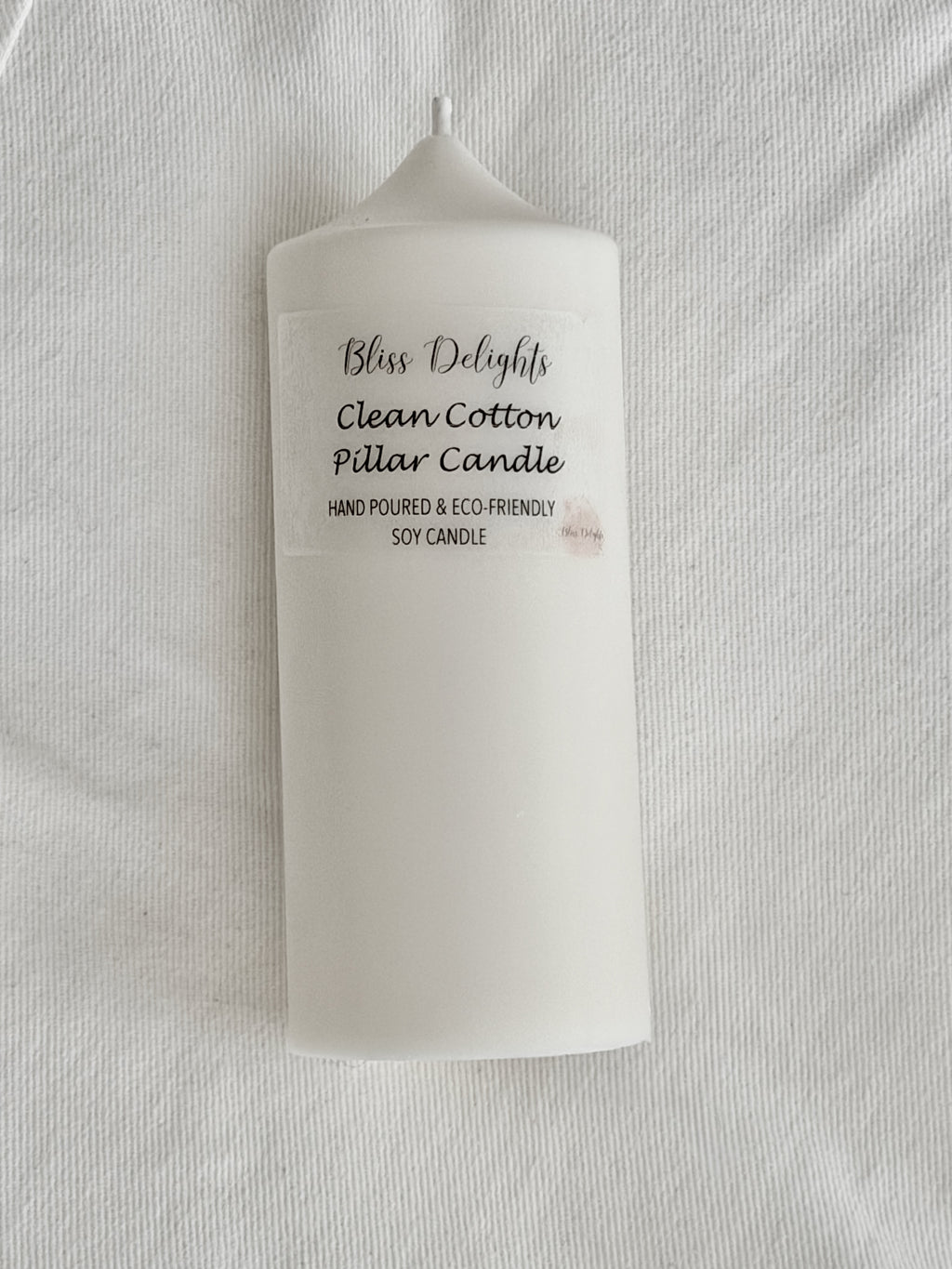 White soy wax pillar candle by Bliss Delights, featuring a Clean Cotton label, photographed against a white textured fabric background.