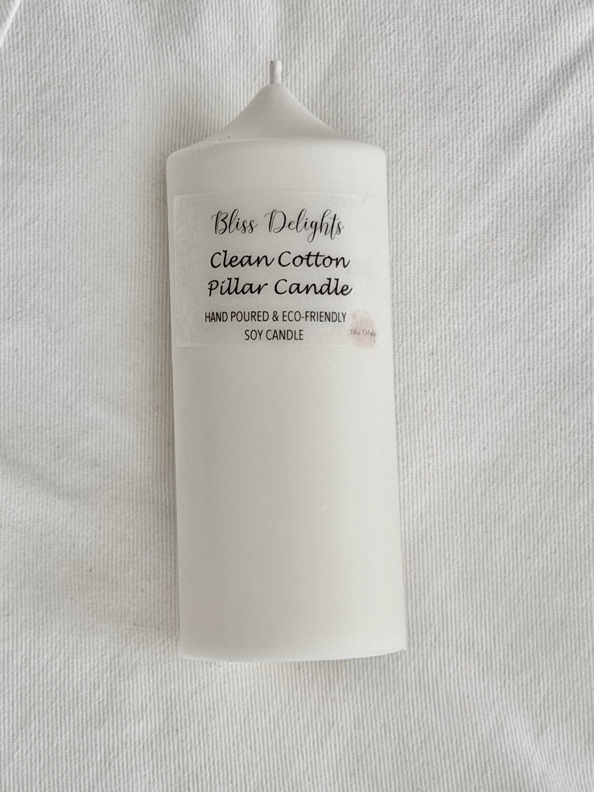 White soy wax pillar candle by Bliss Delights, featuring a Clean Cotton label, photographed against a white textured fabric background.