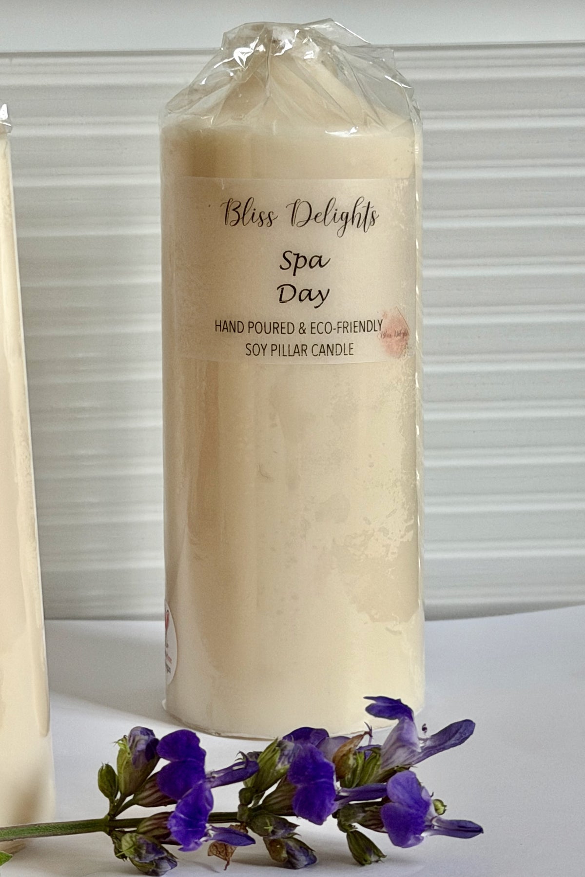 Bliss Delights Spa Day medium soy wax pillar candle in ivory, wrapped in clear packaging with a branded label and displayed beside purple flowers on a white surface.