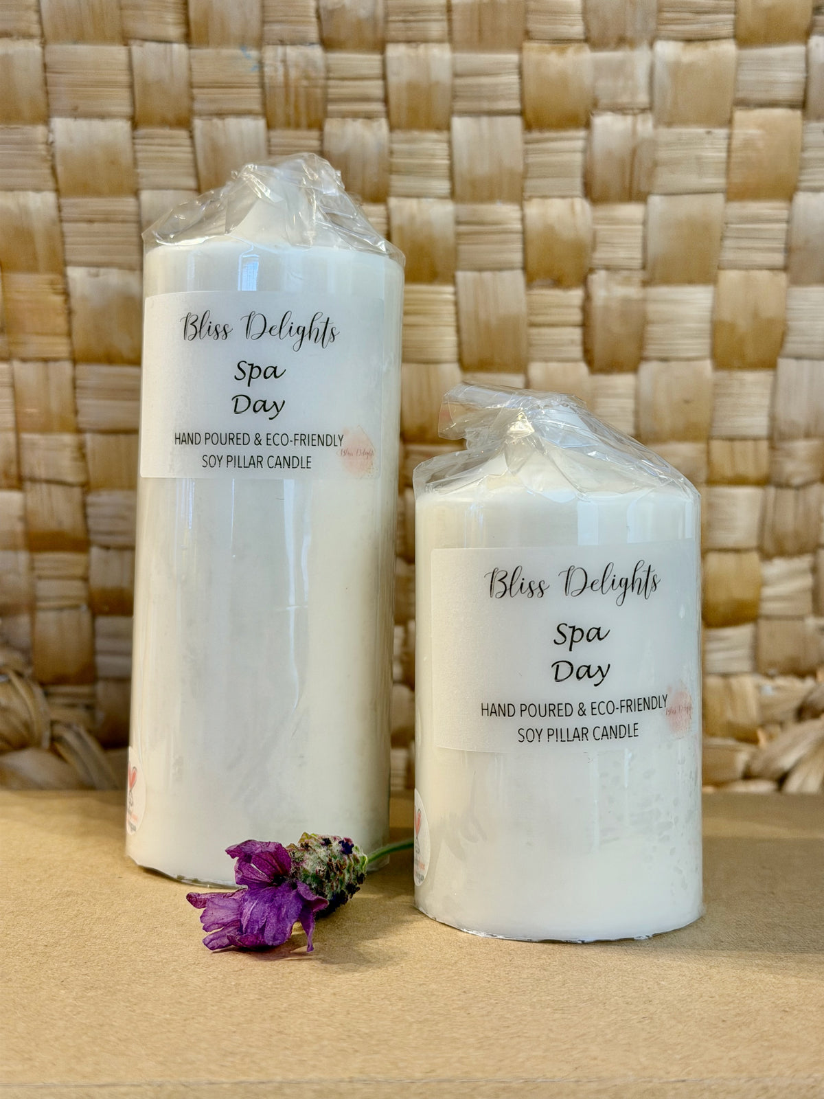 Bliss Delights Spa Day soy wax pillar candles in two sizes, medium and small, displayed in clear packaging against a natural woven background with a purple flower. Handmade ivory pillar candles for home décor or gifting.