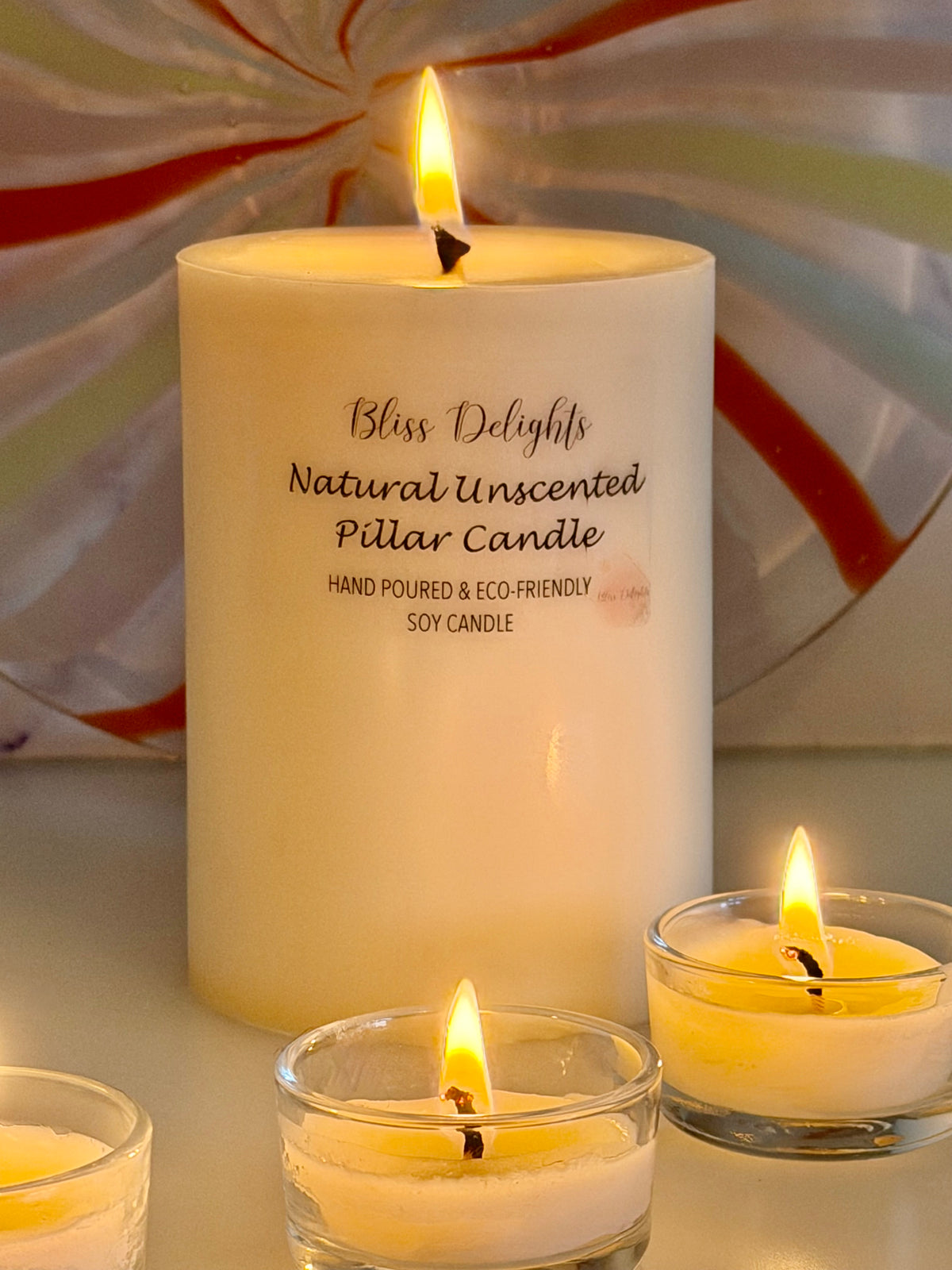 Medium ivory unscented soy pillar candle by Bliss Delights, lit to show its warm glow, with two burning soy tealights in clear glass holders in the foreground against a decorative marble-effect backdrop.