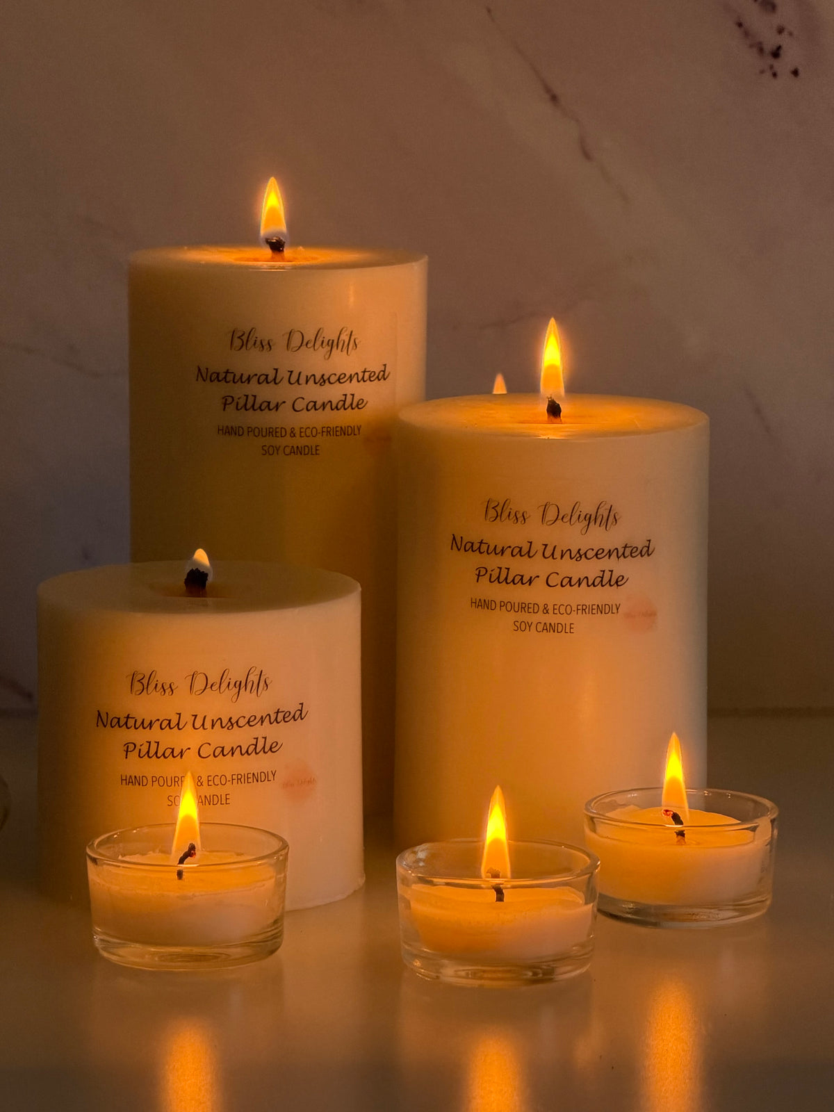Three lit ivory unscented soy pillar candles in small, medium and large sizes, displayed alongside three glowing tealights in glass holders against a marble-effect background in soft candlelight.