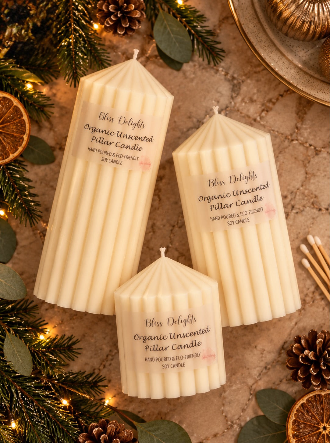 Three Bliss Delights ivory ribbed soy pillar candles, in three different sizes, arranged on a beige marble surface in a sophisticated overhead Christmas flat lay.
