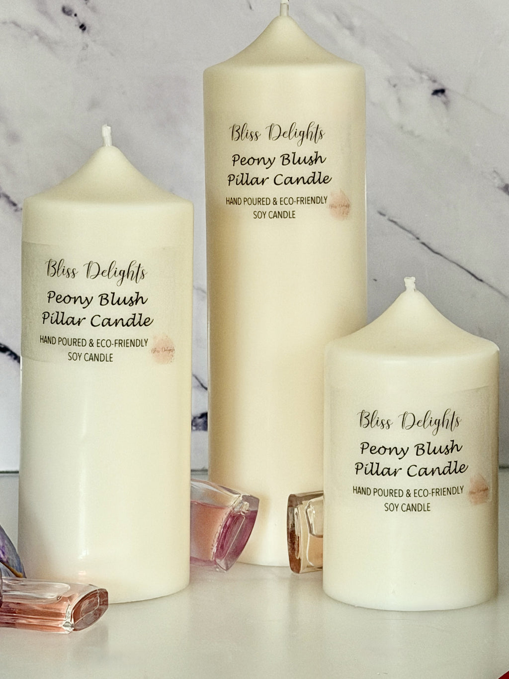 Three cream-coloured Bliss Delights Peony Blush soy wax pillar candles in small, medium and large sizes, arranged against a white marble background with pink and clear perfume bottles as decorative accents.