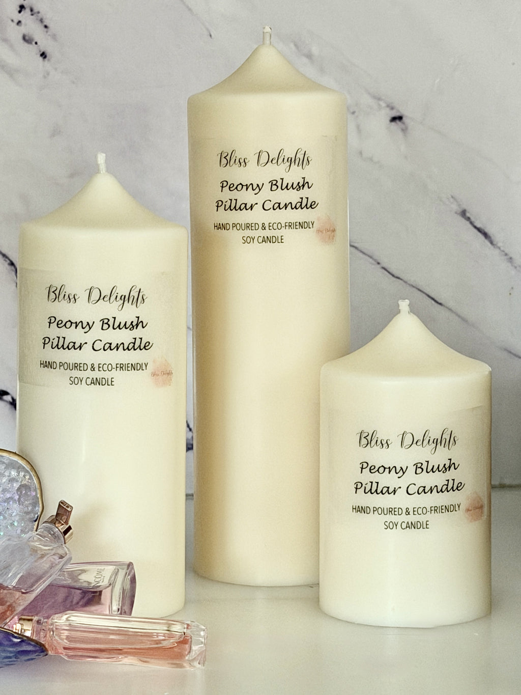 Three cream-coloured Bliss Delights Peony Blush soy wax pillar candles in small, medium and large sizes, displayed against a white marble background with pink perfume bottles and elegant glass fragrance accessories.