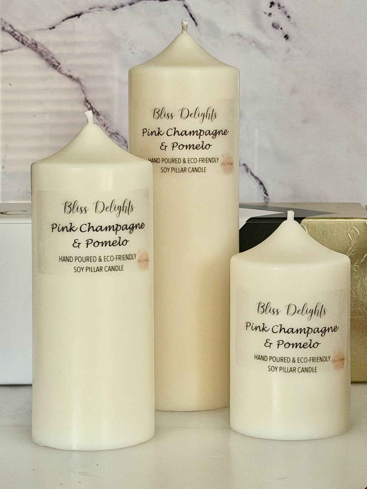 Three cream-coloured Bliss Delights Pink Champagne & Pomelo soy wax pillar candles in small, medium and large sizes, arranged against a white marble background with gold gift packaging behind them.