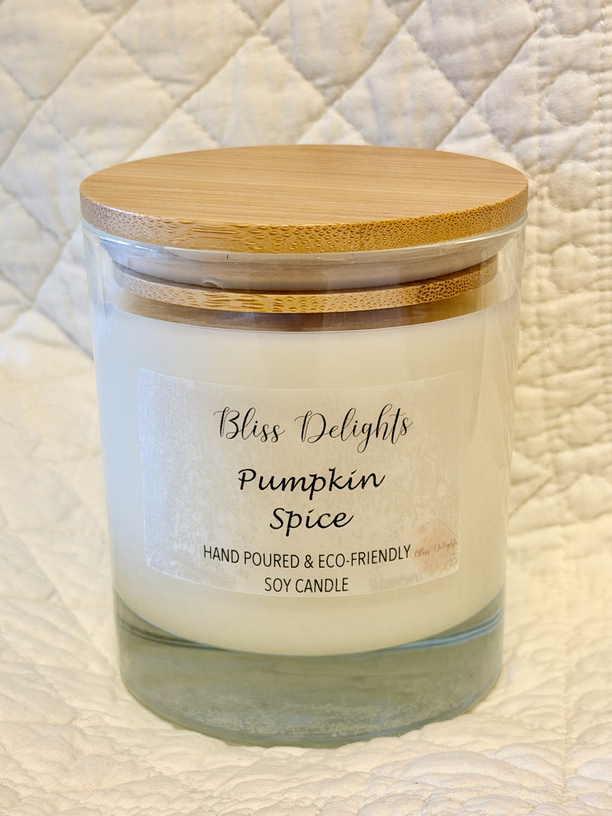Bliss Delights Pumpkin Spice soy candle in a clear glass jar with a natural bamboo lid, displayed against a cream quilted background.