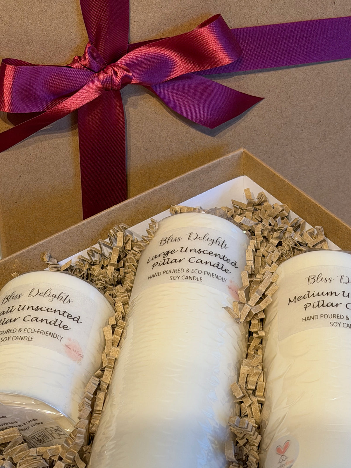 Close-up of three handmade unscented soy pillar candles in small, medium and large sizes, nestled in biodegradable shredded kraft paper inside a gift box with a burgundy satin ribbon.