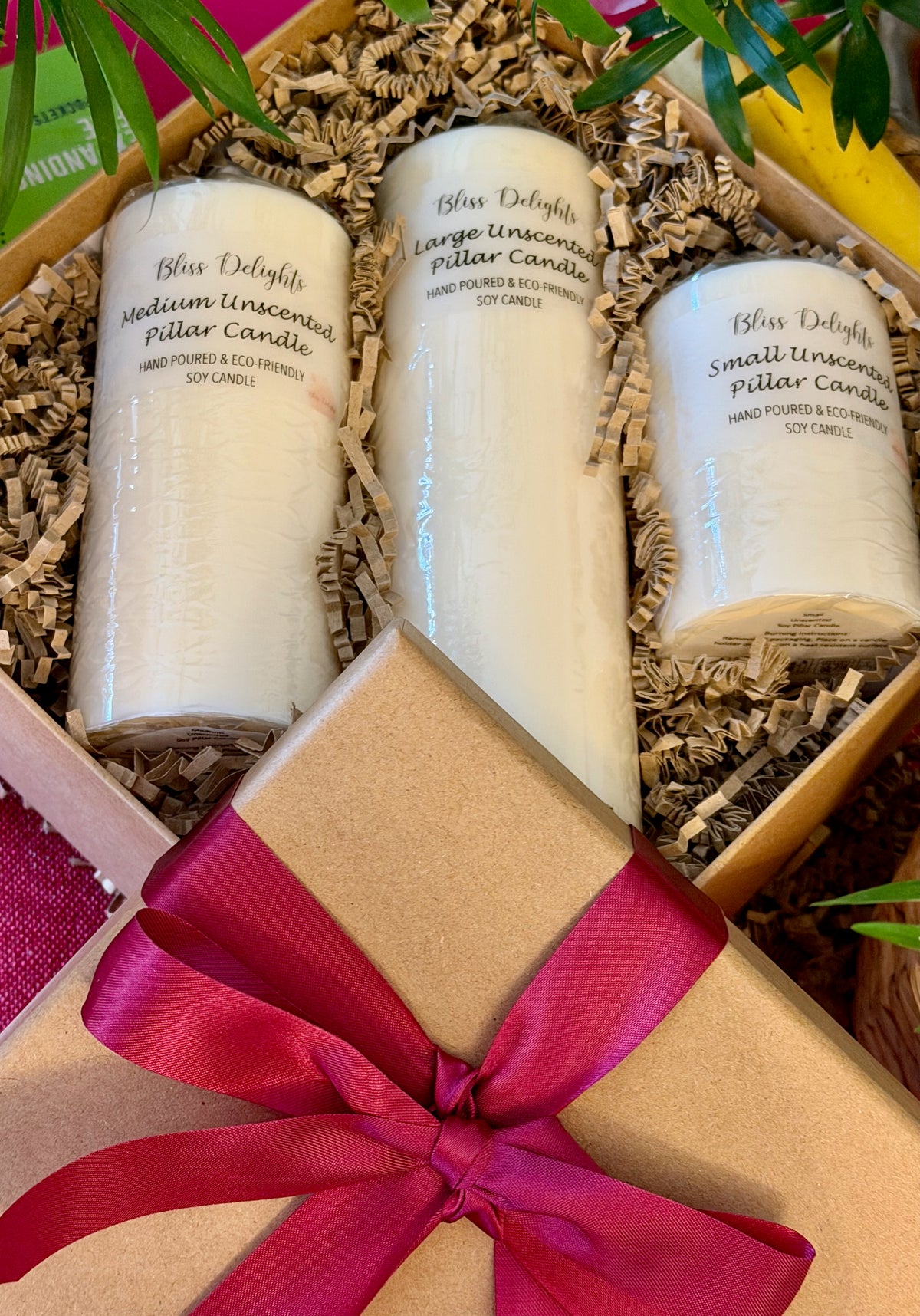 Three unscented ivory soy pillar candles in small, medium and large sizes, presented in a kraft gift box with shredded paper packaging and a burgundy satin ribbon, ready for gifting.