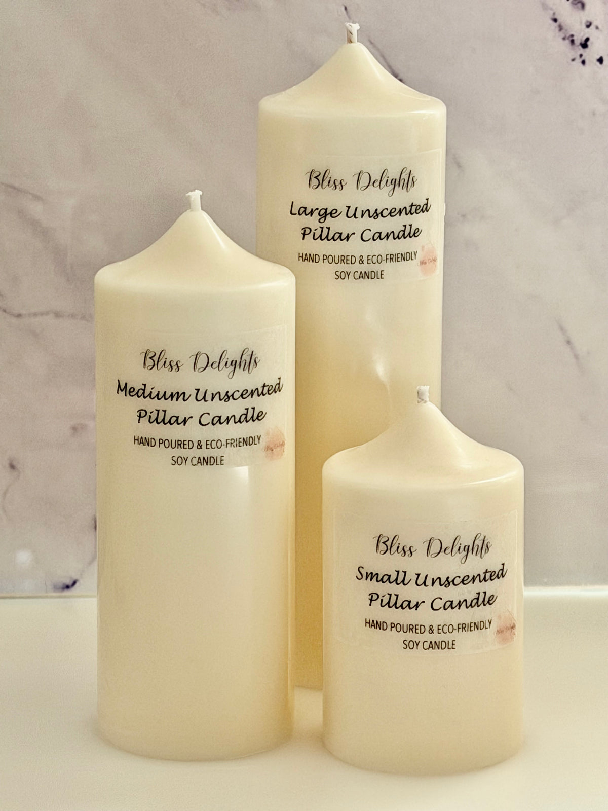 Bliss Delights set of three unscented ivory soy wax pillar candles in small, medium and large sizes, displayed against a light marble background with branded labels. Hand-poured vegan candles for home décor.