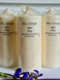Three ivory Bliss Delights Spa Day soy wax pillar candles displayed side by side, wrapped in clear packaging with branded labels. Handmade pillar candles photographed against a light background with purple lavender flowers.