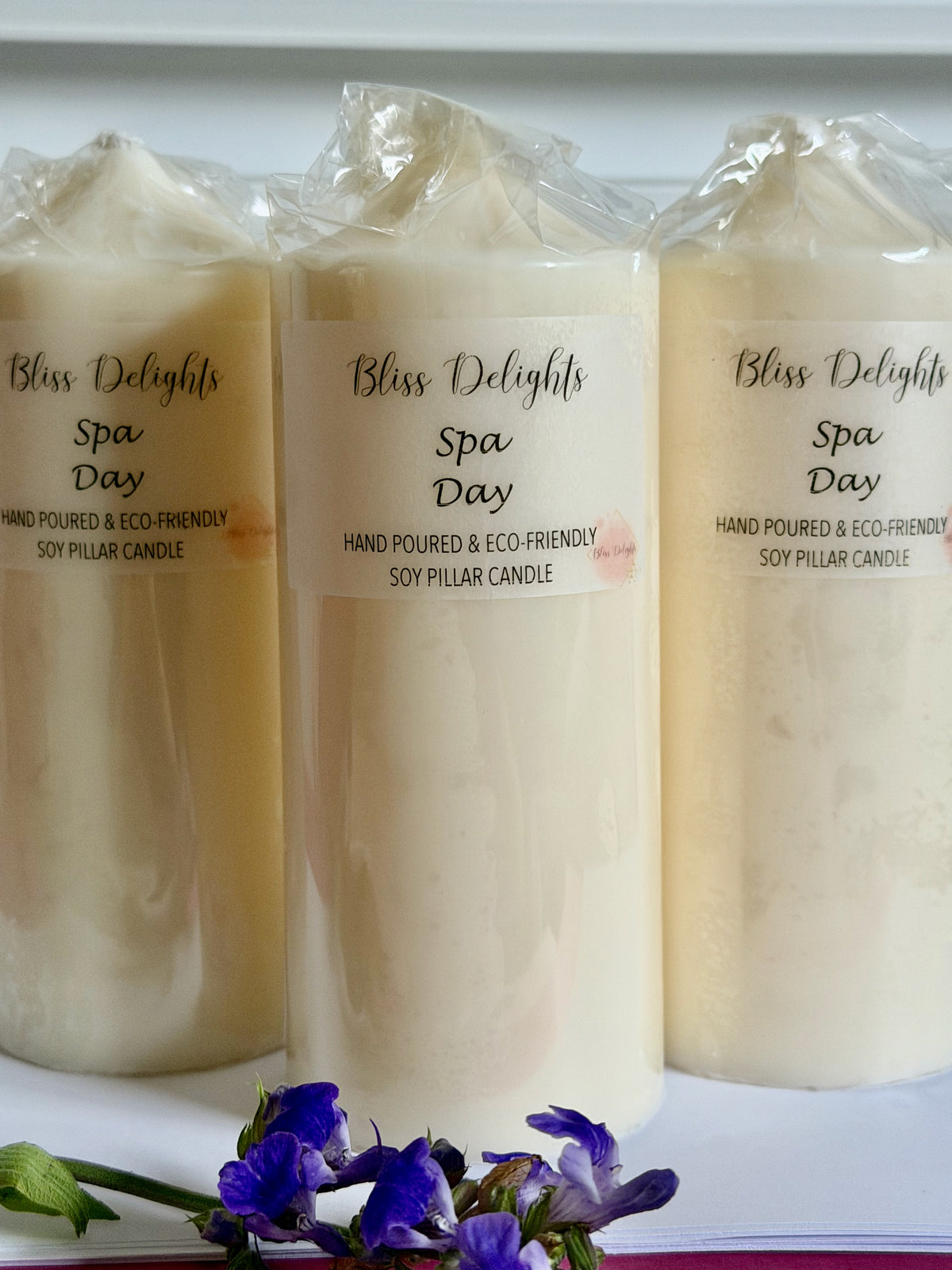 Three ivory Bliss Delights Spa Day soy wax pillar candles displayed side by side, wrapped in clear packaging with branded labels. Handmade pillar candles photographed against a light background with purple lavender flowers.