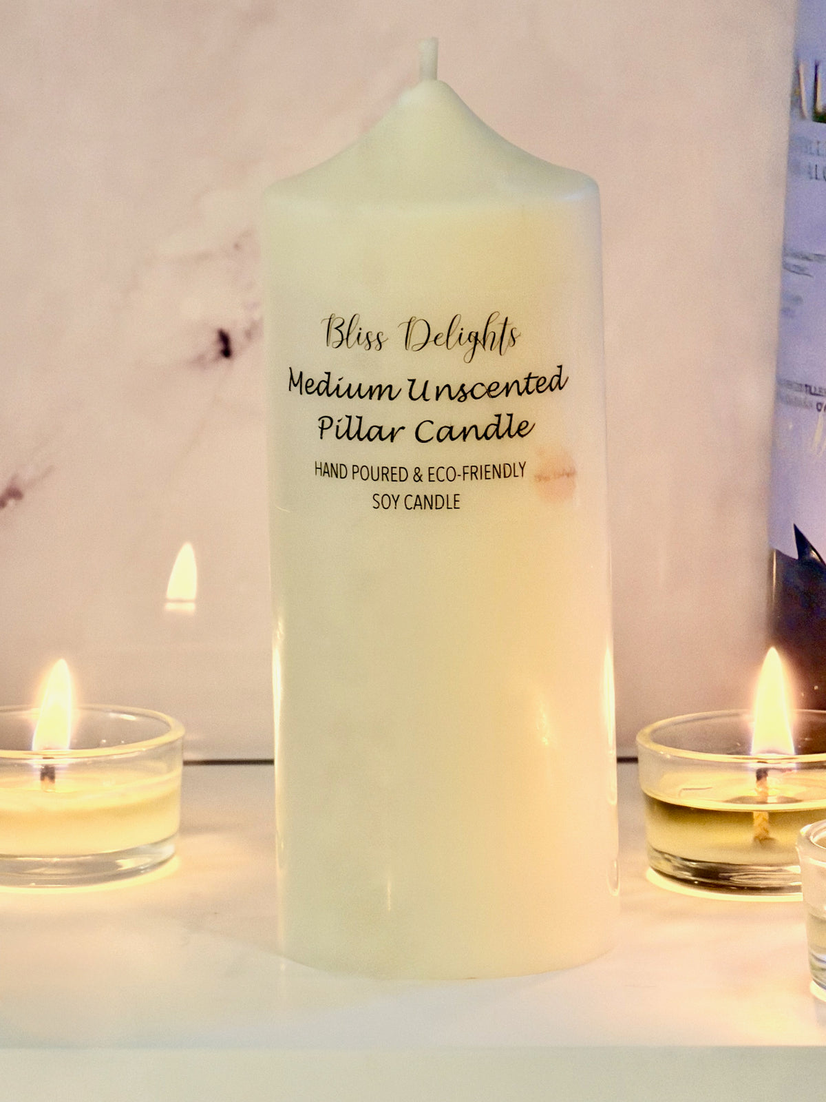 Medium ivory soy wax pillar candle by Bliss Delights, with a minimalist label, styled alongside lit tealights on a white surface.