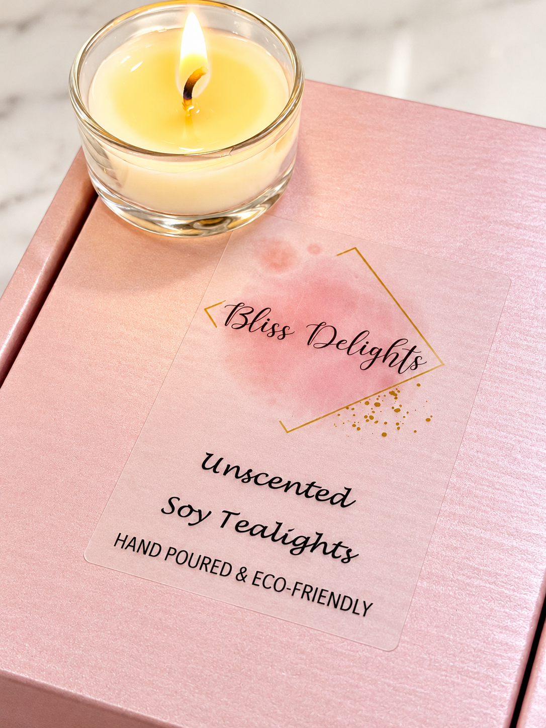 Bliss Delights unscented soy tealights in a clear glass holder, displayed on a pink gift box with a transparent label, styled on a light marble background.