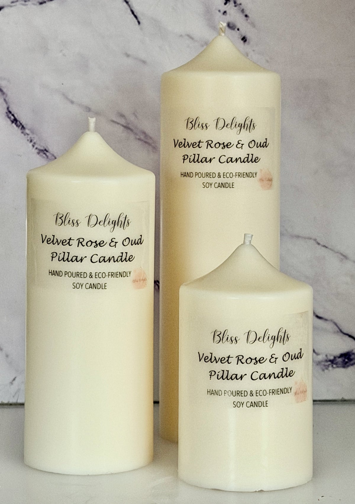 Three cream-coloured Bliss Delights Velvet Rose & Oud soy wax pillar candles in small, medium and large sizes. The candles are arranged against a white marble background.