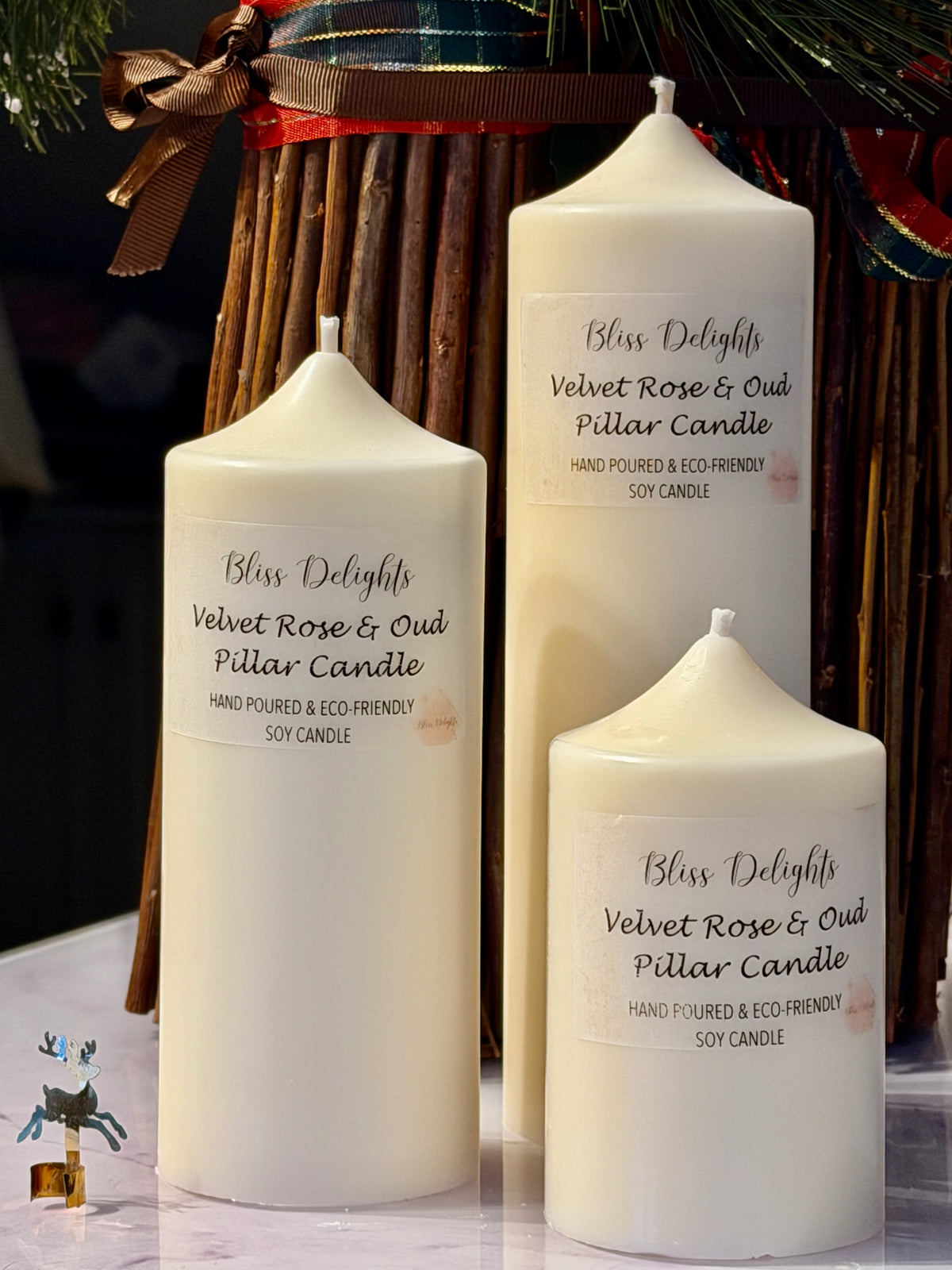 Three cream-coloured Bliss Delights Velvet Rose & Oud soy wax pillar candles in small, medium and large sizes. The candles are arranged against a white marble background.
